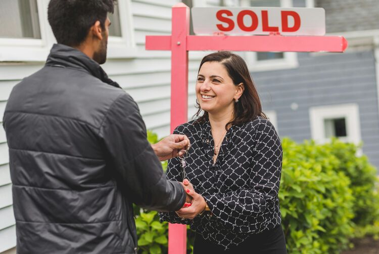 Real estate agent handing house keys to a new homeowner in front of a sold sign.